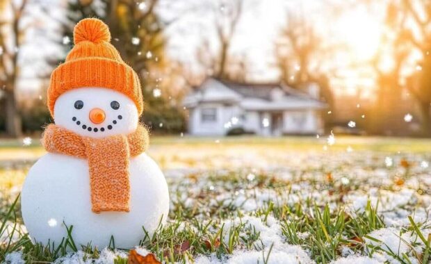 cheerful snowman with an orange hat and scarf stands on a lawn covered in snow and fallen leaves with a house in the background